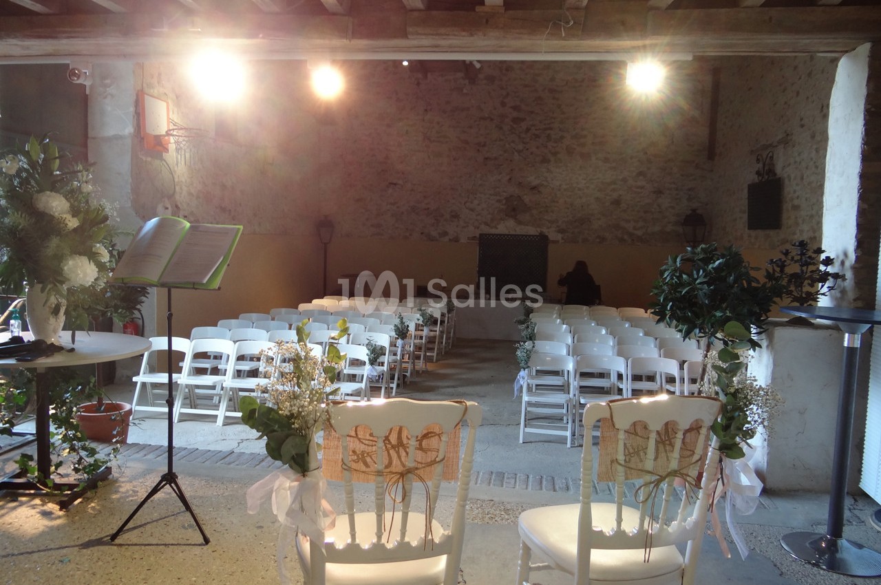 Salle décorée pour une cérémonie, avec chaises blanches alignées, fleurs et lumière douce sous une charpente en bois.