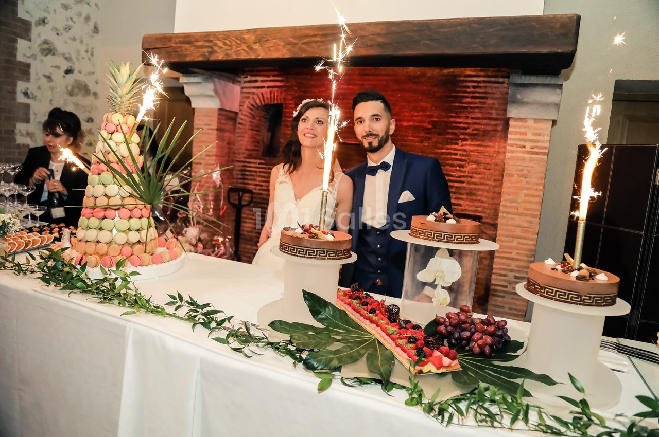 Un couple souriant devant une table décorée avec des desserts, des macarons et des gâteaux ornés de cierges magiques.