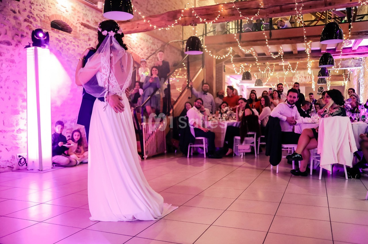 Un couple de mariés danse sous des lumières féeriques, entouré d'invités dans une salle décorée.