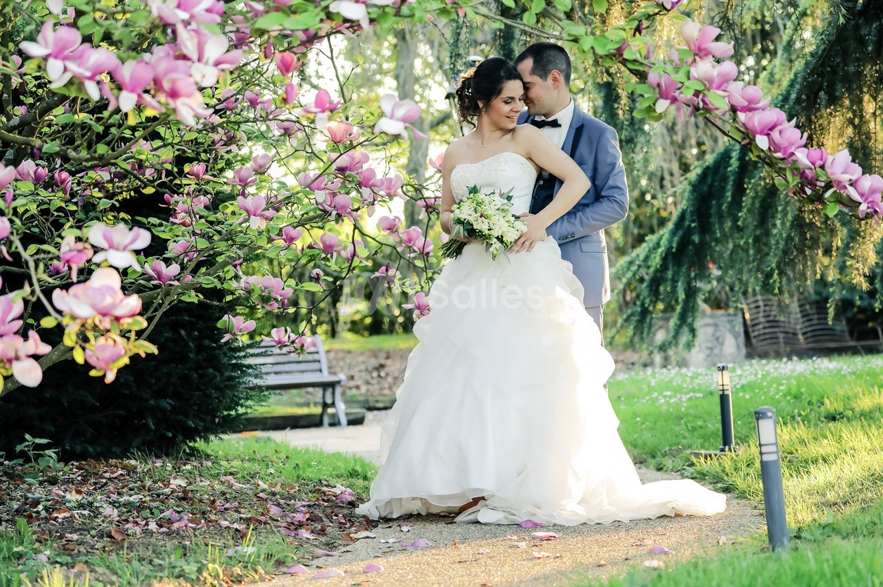 Un couple de mariés pose dans un jardin fleuri, entouré de magnolias roses et d'une lumière douce.
