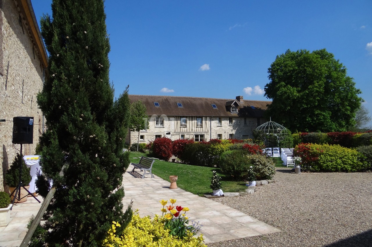 Cour d'une propriété en pierre avec jardin fleuri, allée gravillonnée, et tonnelle sous un ciel bleu.