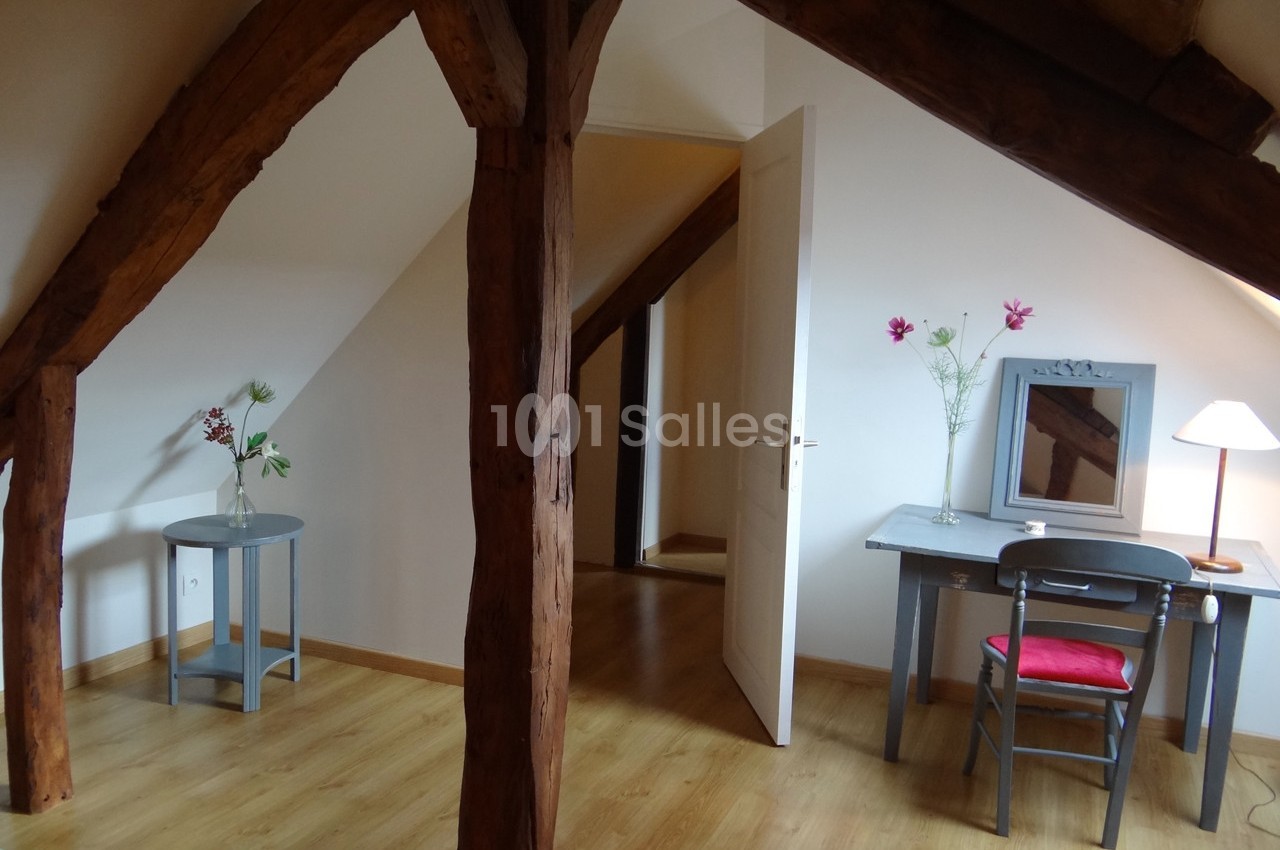 Pièce mansardée avec poutres apparentes, un bureau, une chaise, une lampe et des fleurs sur des meubles en bois.