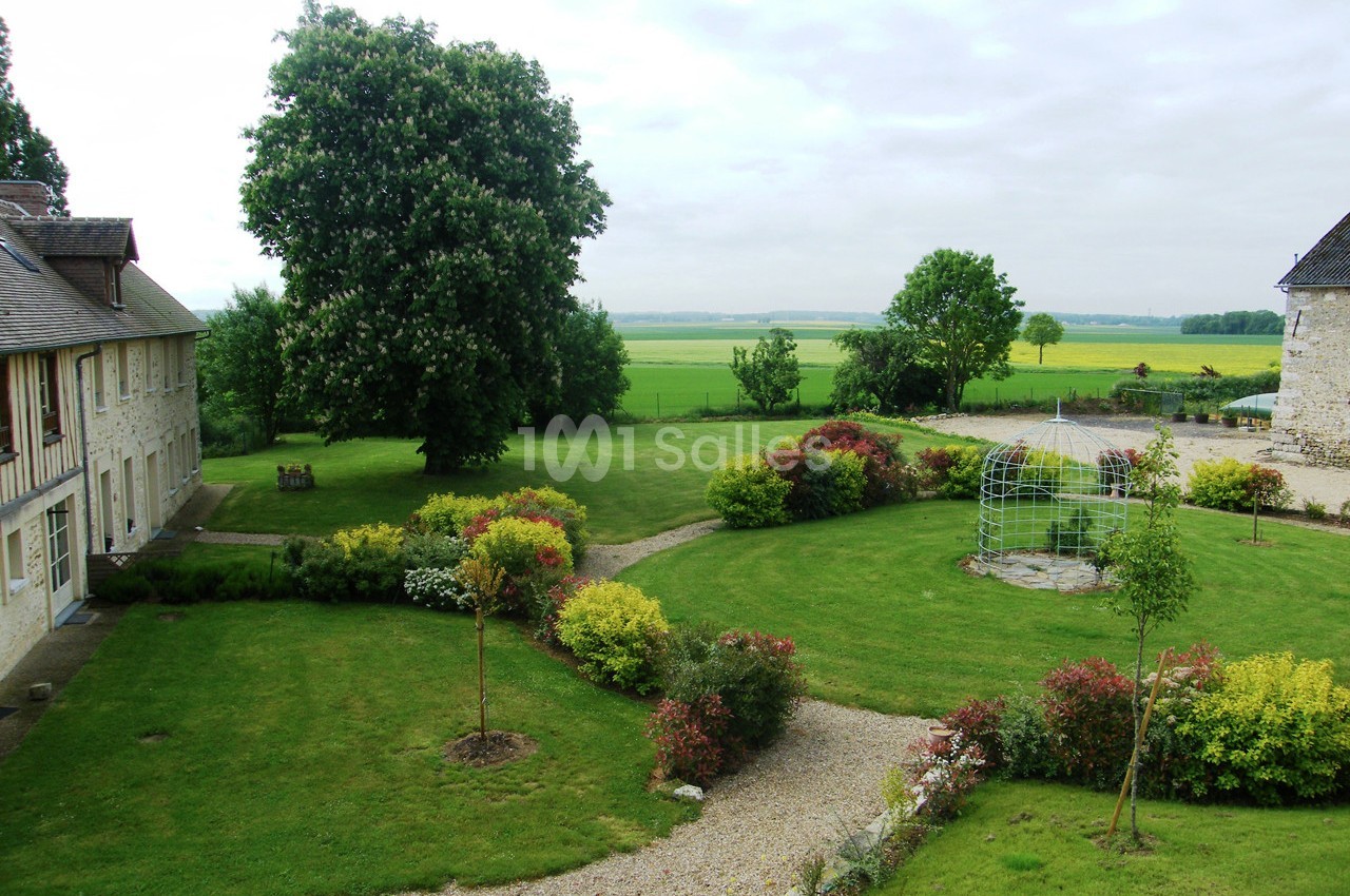 Jardin paysager avec pelouse, massifs fleuris, allées gravillonnées, tonnelle métallique et bâtiments en pierre.