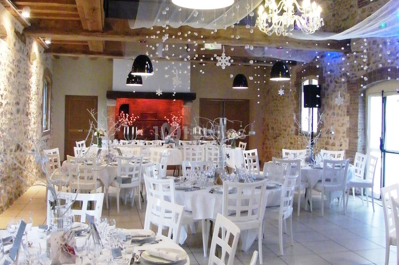 Salle de réception décorée pour un événement, avec tables rondes, chaises blanches et ornements suspendus.
