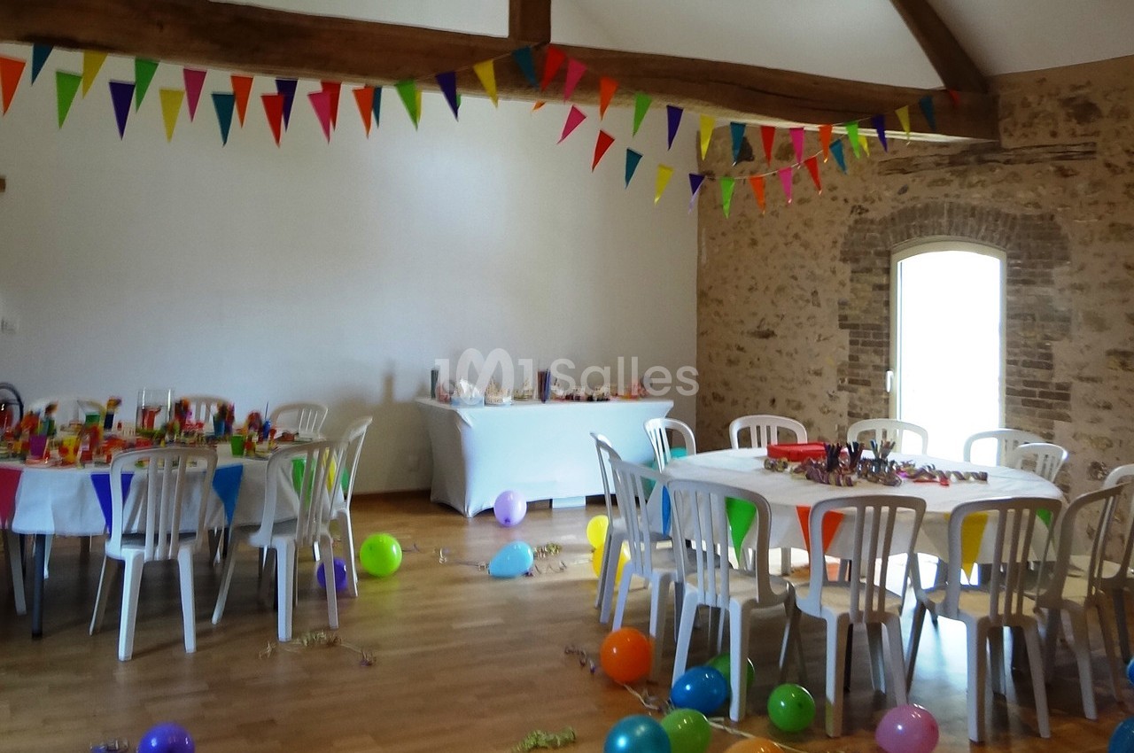 Salle décorée pour une fête avec des tables dressées, des guirlandes colorées et des ballons au sol.