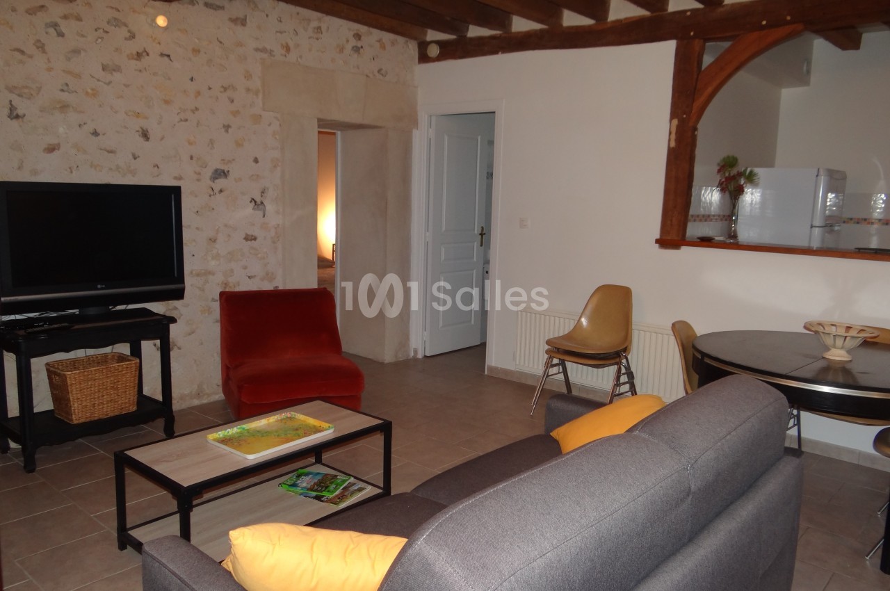 Salon avec murs en pierre, canapé gris, fauteuil rouge, table ronde, télévision et cuisine ouverte en arrière-plan.