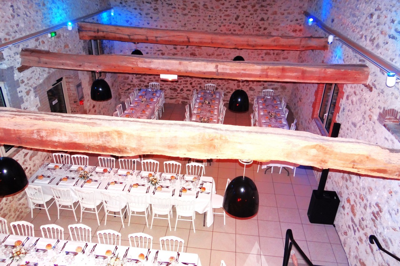 Salle de réception en pierre avec poutres apparentes, tables dressées et chaises blanches alignées pour un événement.