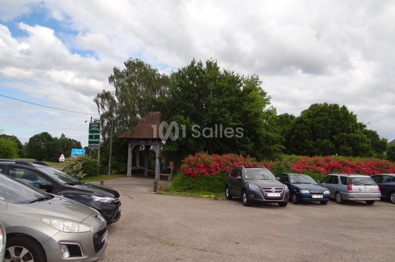 Parking extérieur avec plusieurs voitures, entouré de buissons fleuris et d'arbres sous un ciel partiellement nuageux.
