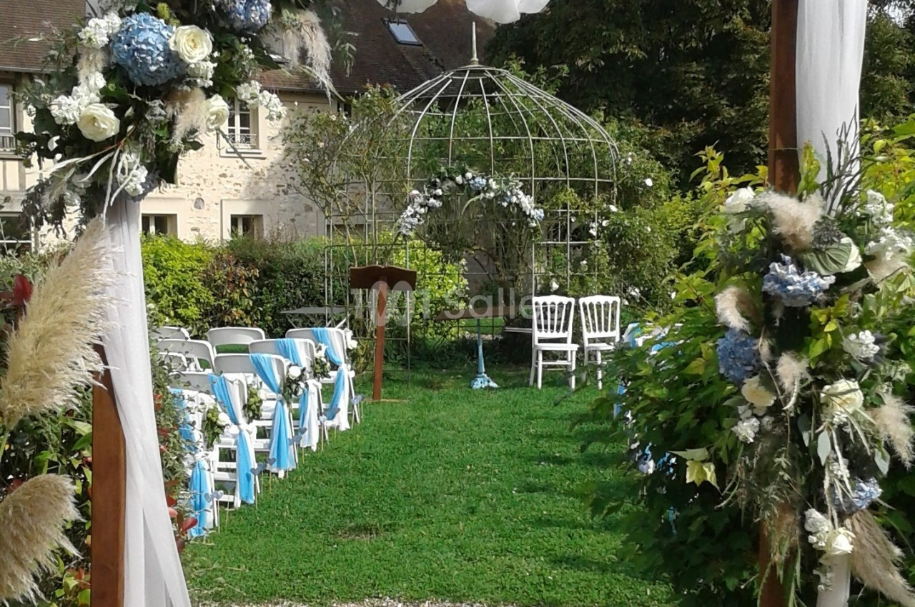 Cérémonie en extérieur avec chaises décorées, arche florale et structure métallique dans un jardin verdoyant.
