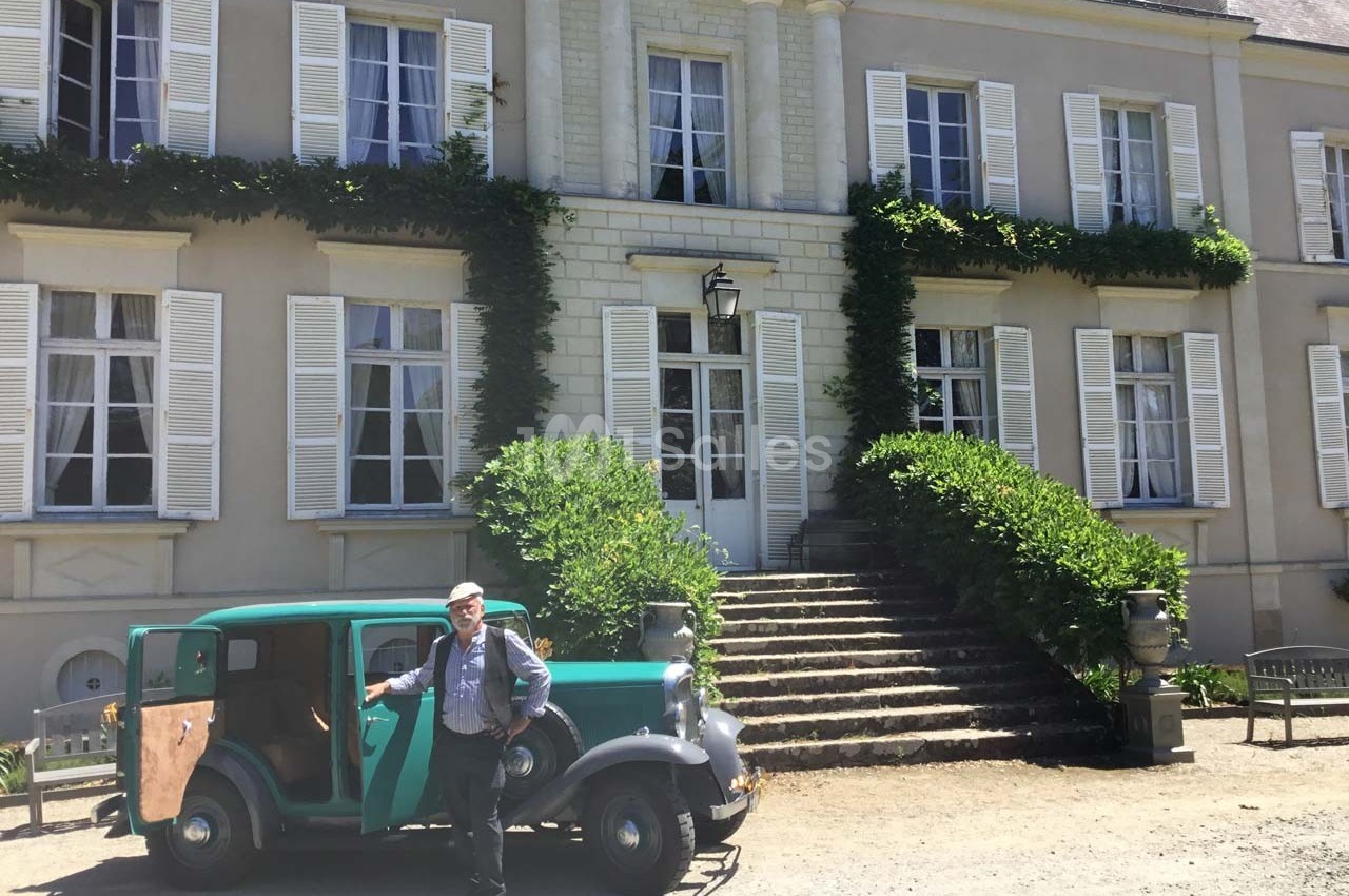 Un homme pose près d'une voiture ancienne devant un bâtiment en pierre avec des volets blancs et des escaliers.