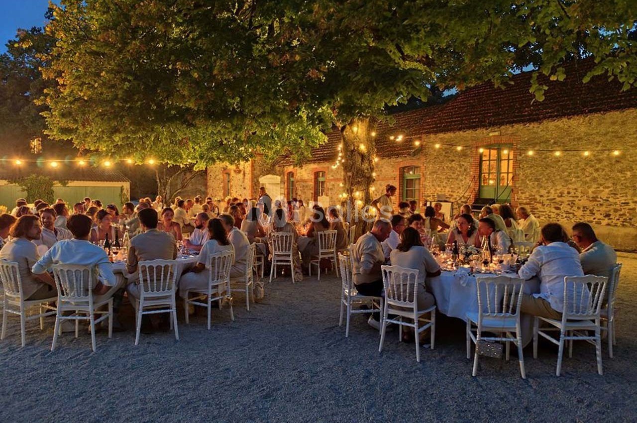 Dîner en plein air sous des guirlandes lumineuses, avec des invités assis autour de tables rondes près d'un bâtiment en…