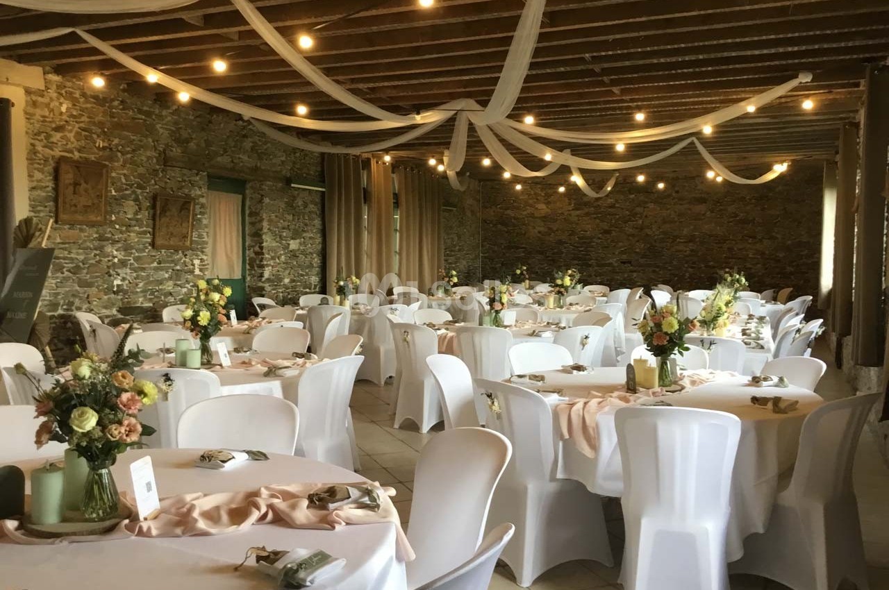 Salle de réception décorée avec des tables rondes, nappes blanches, bouquets de fleurs et guirlandes lumineuses suspendues.