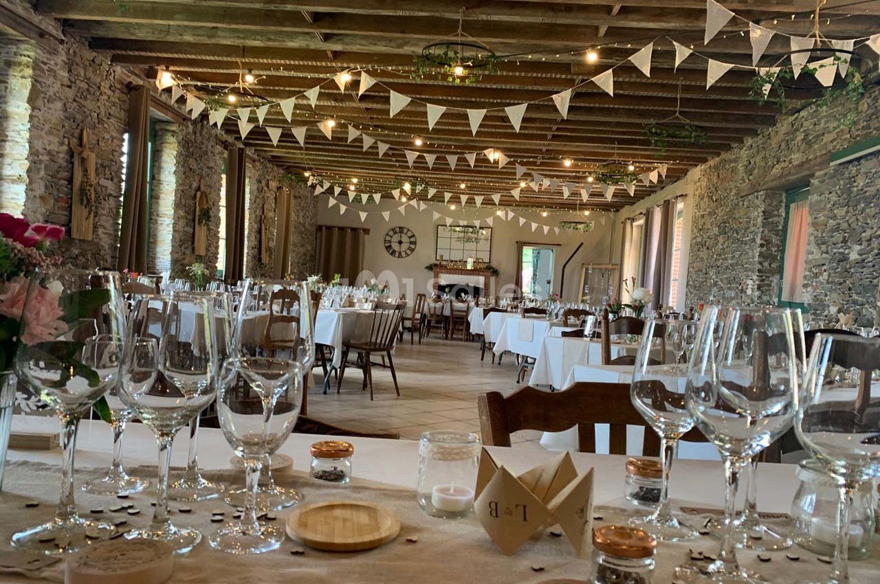 Grande salle de réception rustique décorée de guirlandes, avec tables dressées et murs en pierre apparente.