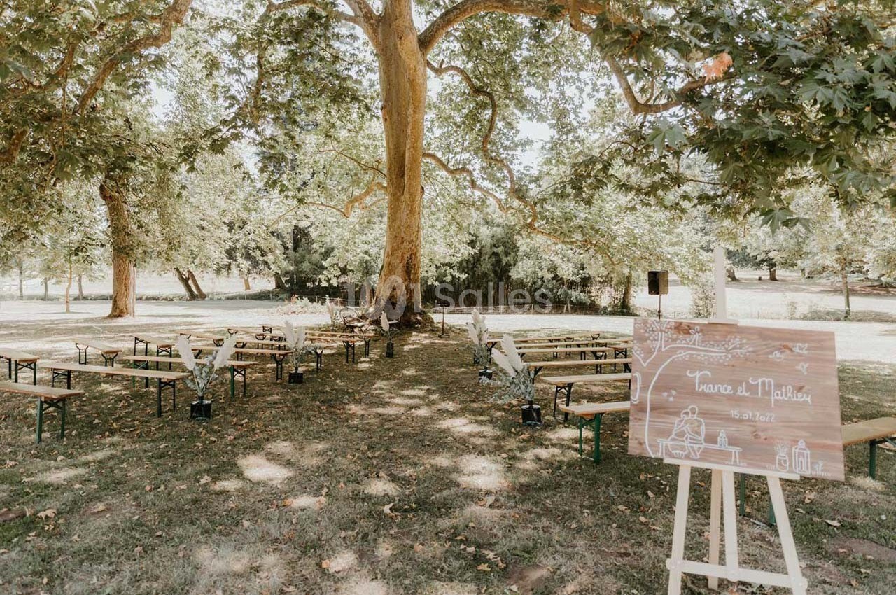 Chaises et bancs disposés en extérieur sous un grand arbre, avec un panneau de mariage en bois au premier plan.