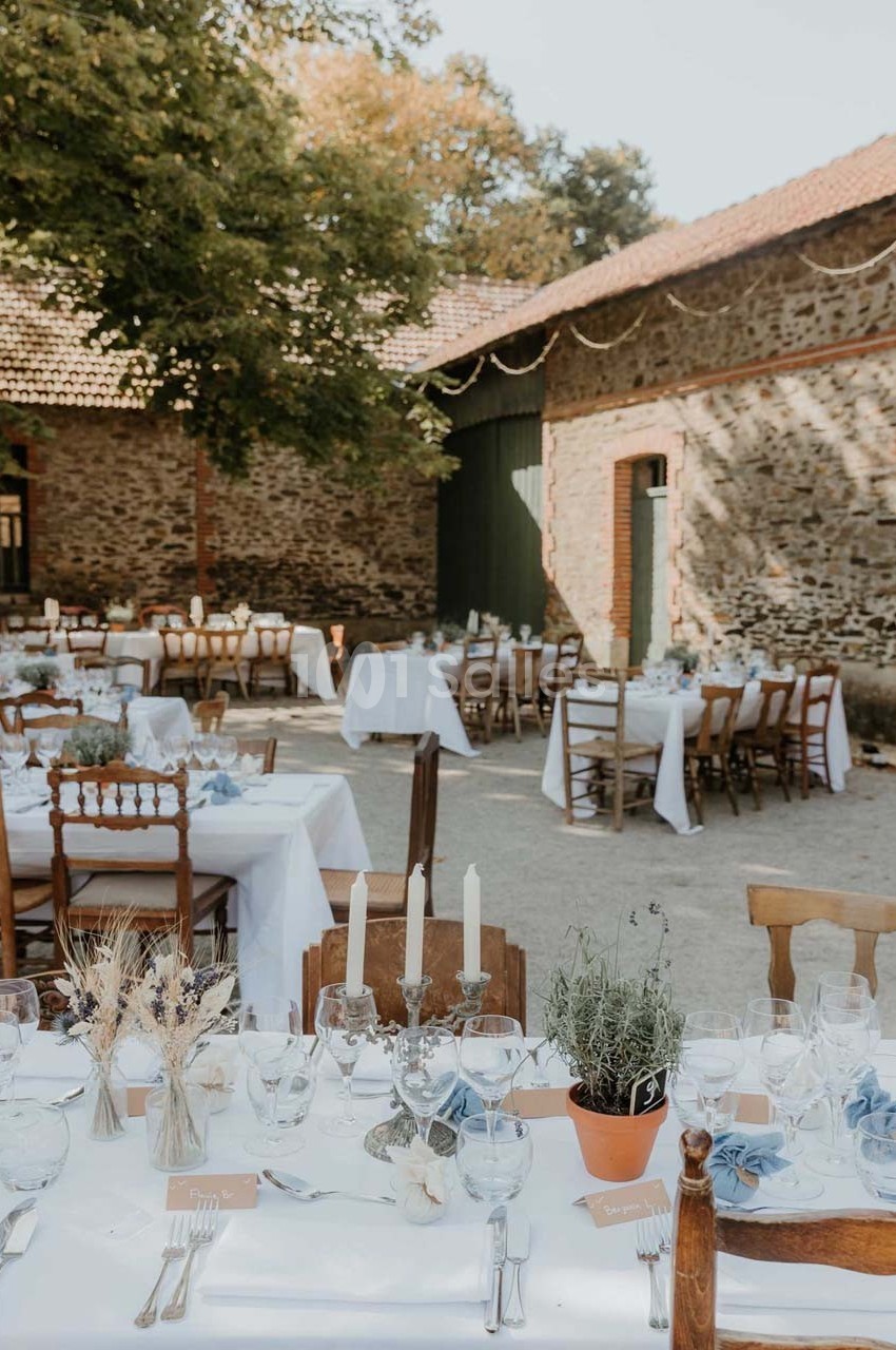 Tables dressées pour un repas en extérieur dans une cour pavée, entourée de bâtiments en pierre et d'arbres.