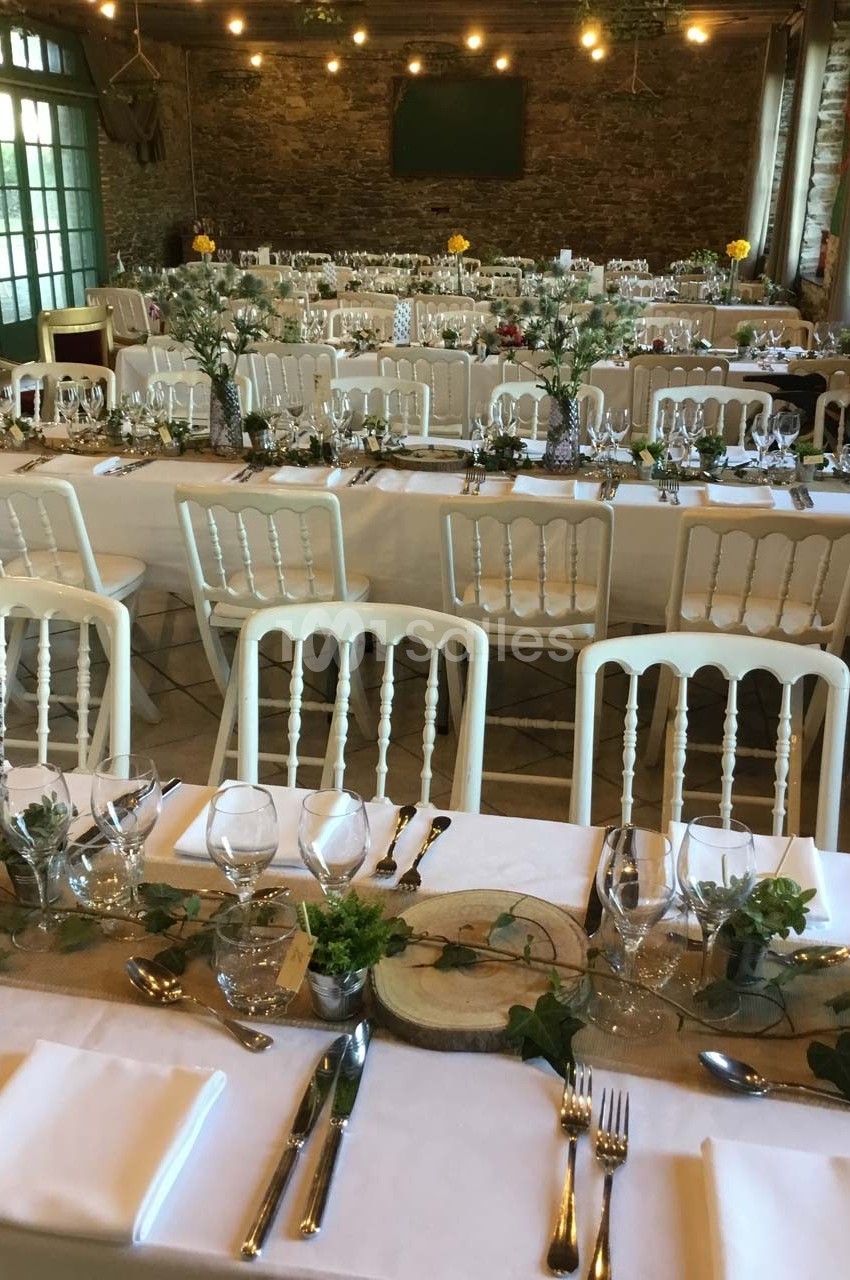 Salle de réception décorée avec des tables alignées, nappes blanches, chaises en bois clair et ornements végétaux.
