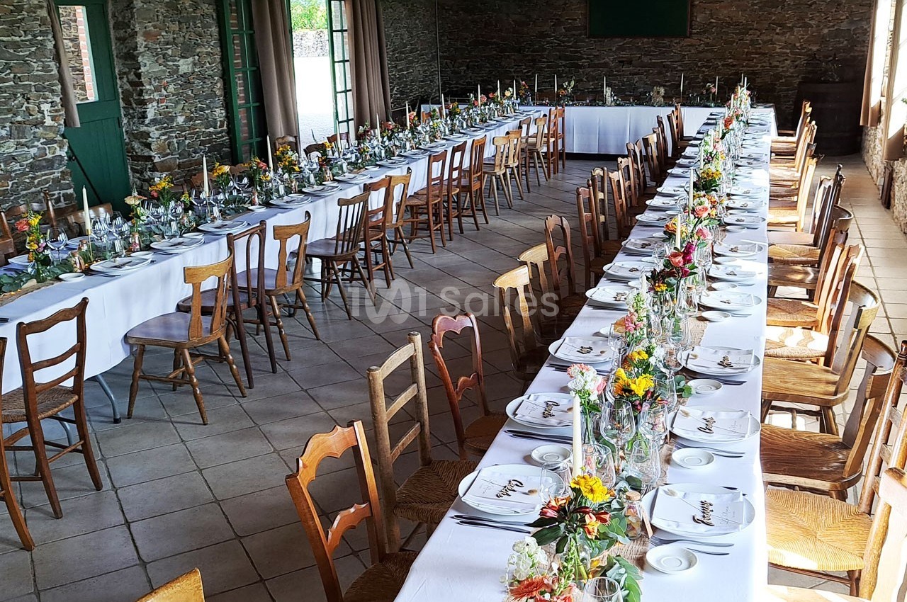 Tables longues dressées avec nappes blanches, vaisselle et décorations florales colorées dans une salle en pierre.