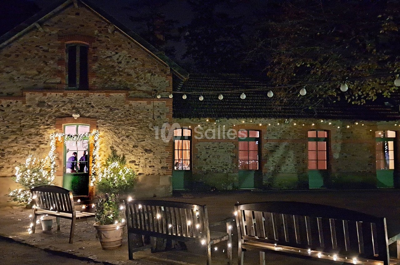 Façade en pierre éclairée de guirlandes lumineuses, avec bancs et végétation décorée dans une ambiance nocturne.