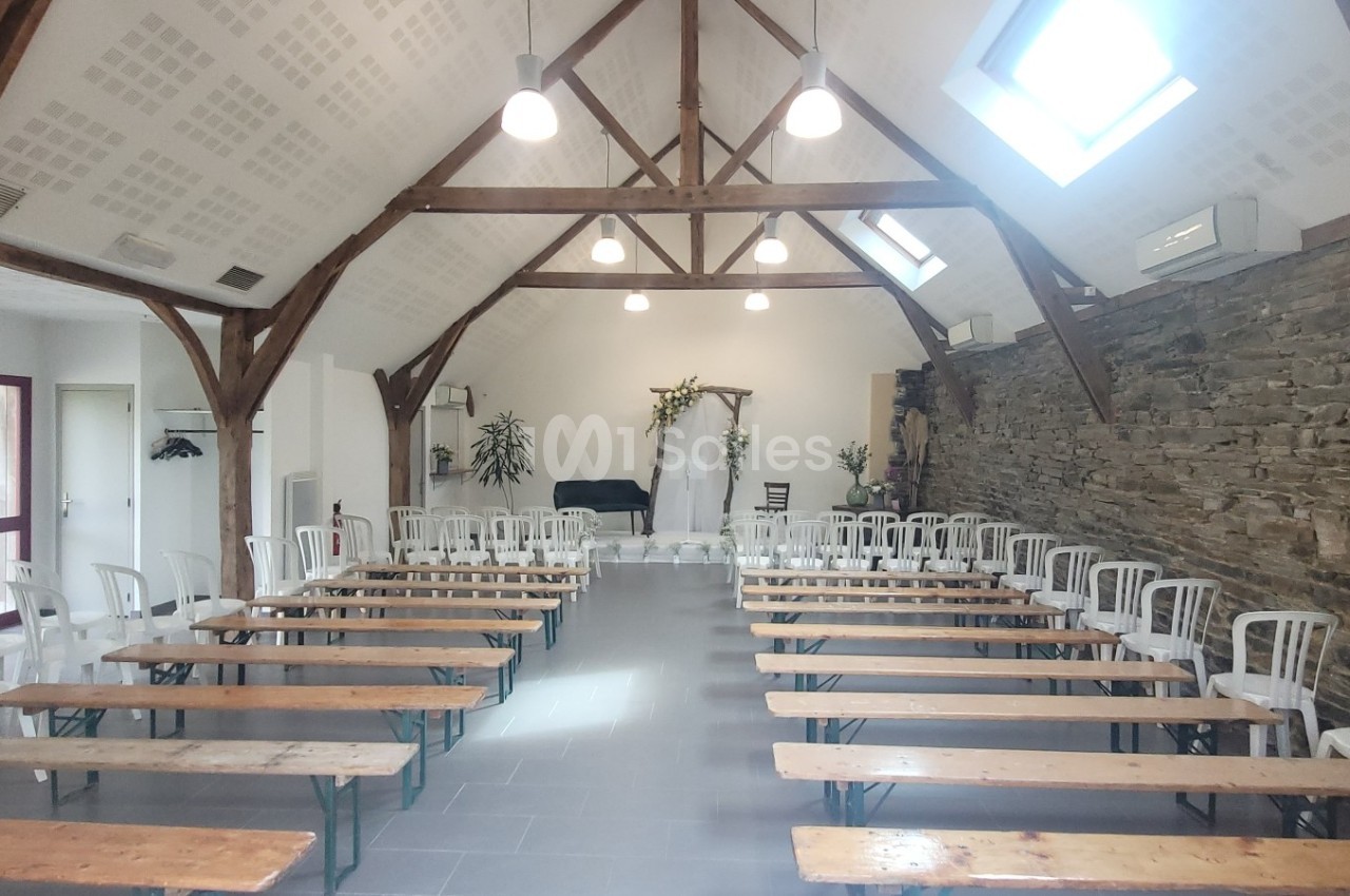 Salle lumineuse avec des bancs en bois et des chaises blanches alignés, décorée pour un événement avec des fleurs.