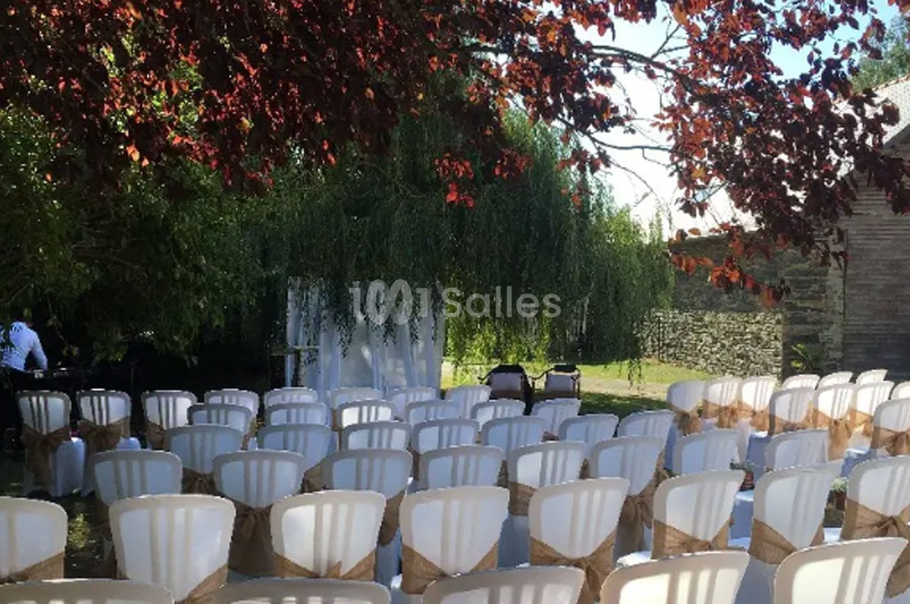 Chaises blanches alignées en extérieur sous des arbres, décorées de rubans beige, prêtes pour une cérémonie.