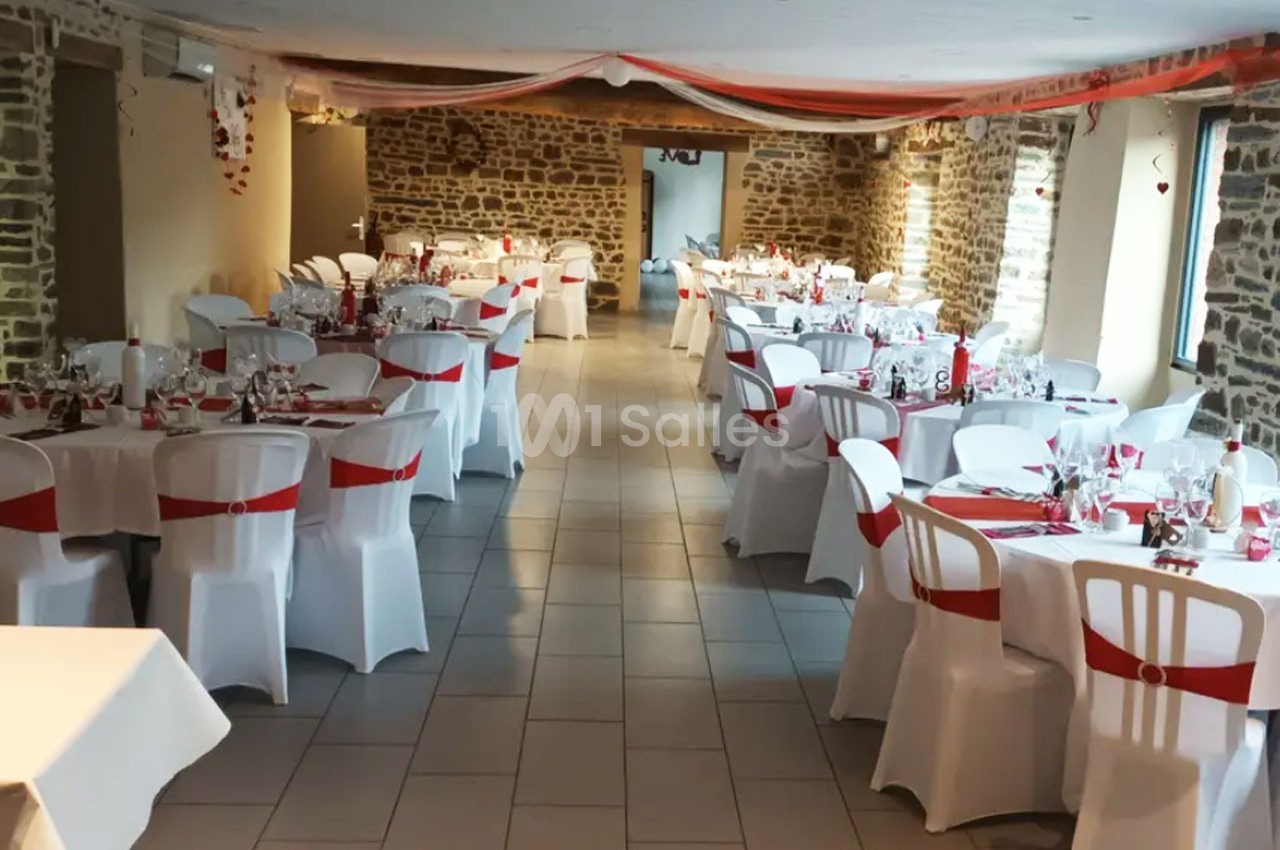 Salle de réception décorée avec des tables rondes, nappes blanches et accents rouges, murs en pierre apparente.