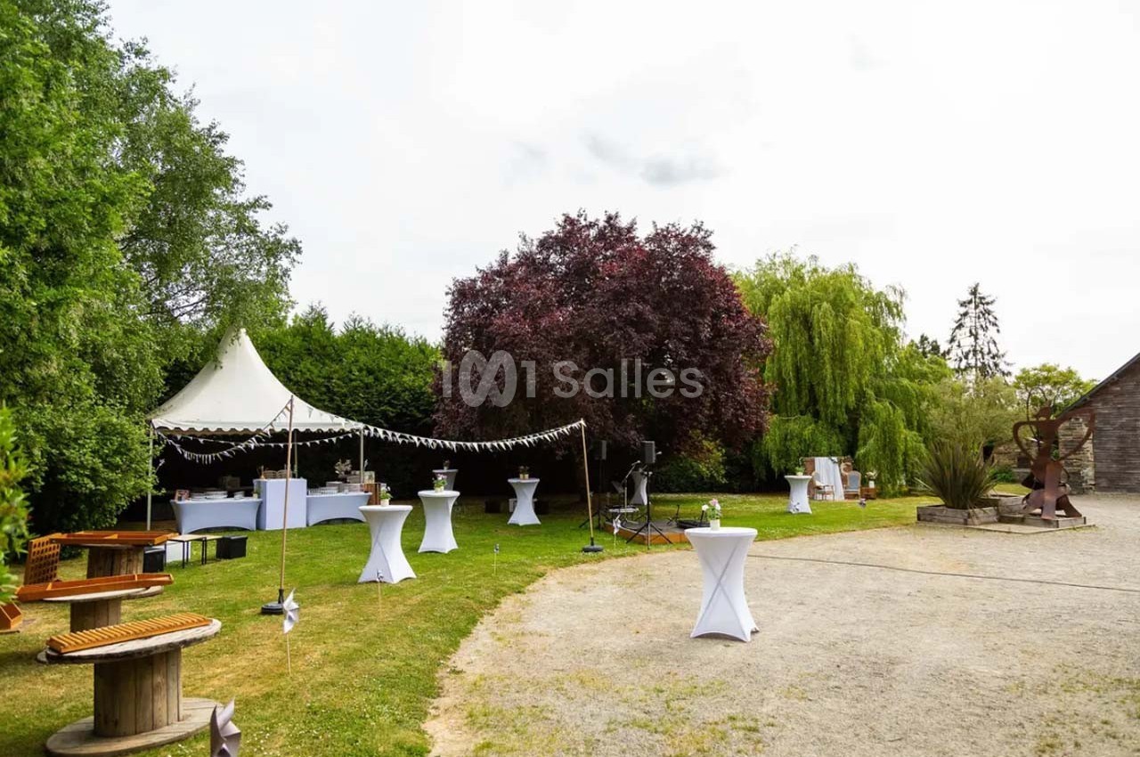 Jardin aménagé pour un événement avec tables hautes, tente blanche et décorations sous des arbres.