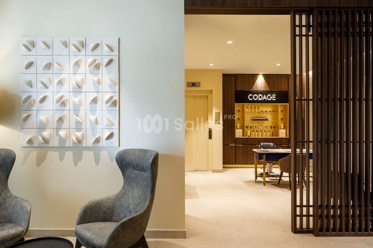 Deux fauteuils gris et une table basse dans un espace lumineux, avec un mur décoratif et un accès à une pièce adjacente.