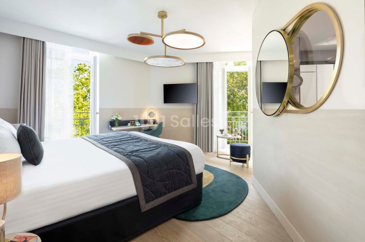 Chambre lumineuse avec lit double, bureau, télévision, miroir rond et balcon donnant sur des arbres.