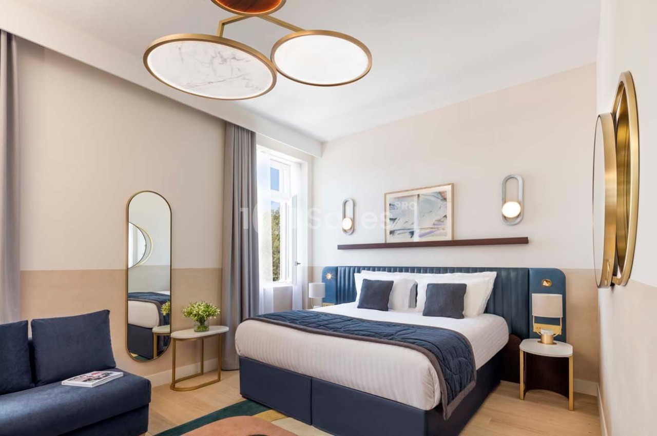 Chambre lumineuse avec lit double, canapé, table basse, miroir, et décoration moderne dans des tons neutres et bleus.