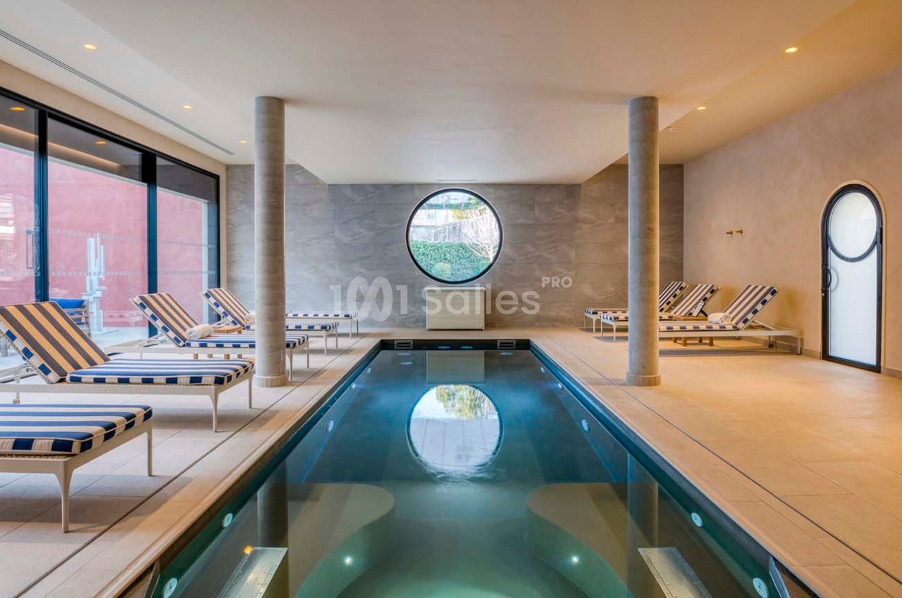 Piscine intérieure avec chaises longues rayées, éclairage doux et grande fenêtre ronde donnant sur l'extérieur.