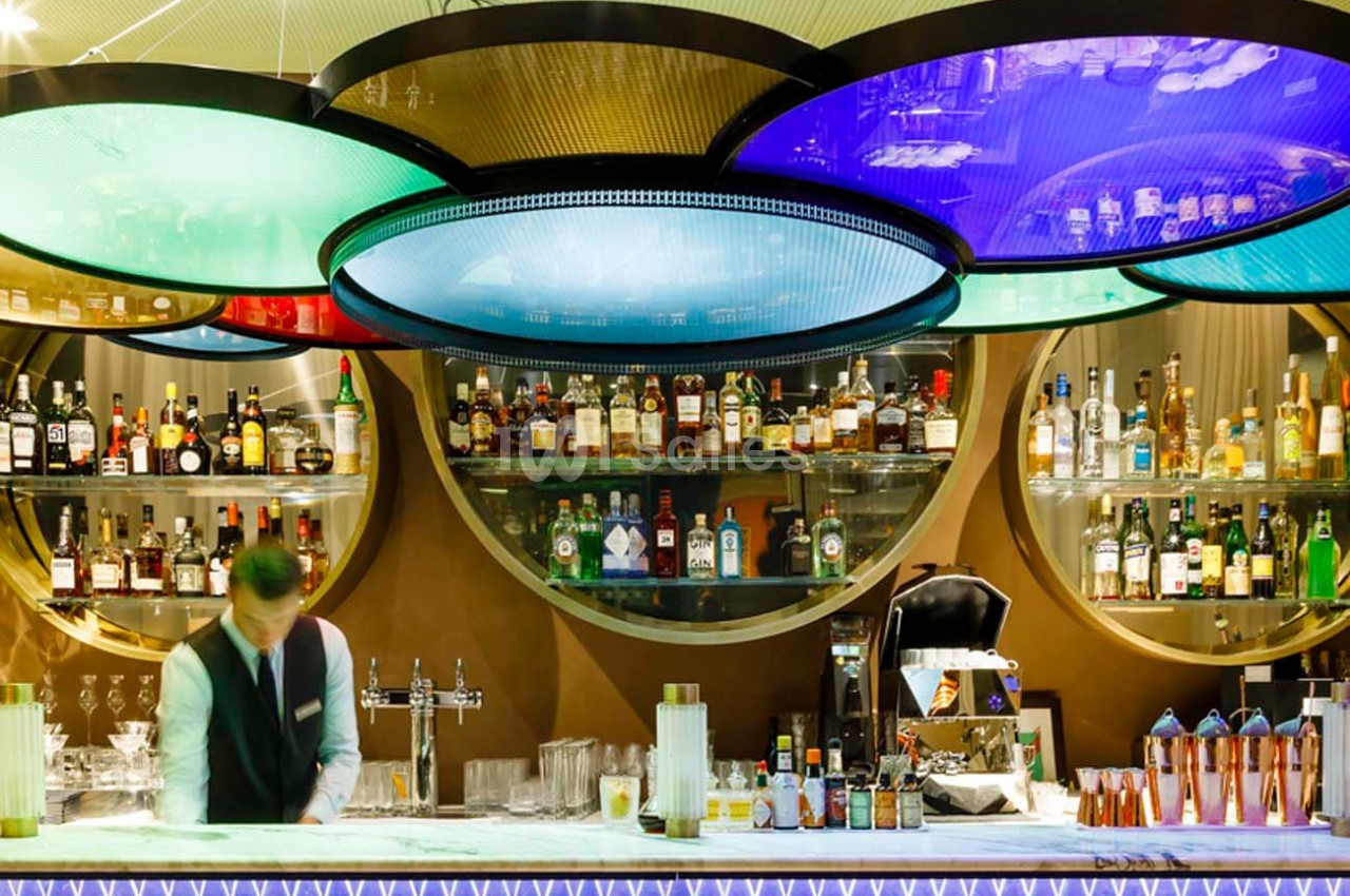 Un barman prépare des boissons derrière un comptoir, avec des étagères remplies de bouteilles et des lumières colorées au…