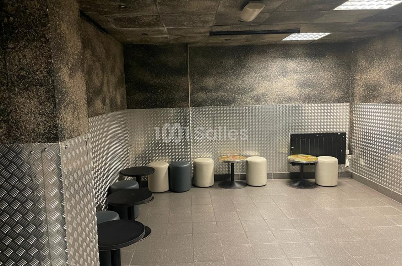 Salle avec murs texturés, sol carrelé, tables noires rondes et tabourets rembourrés disposés contre les murs.