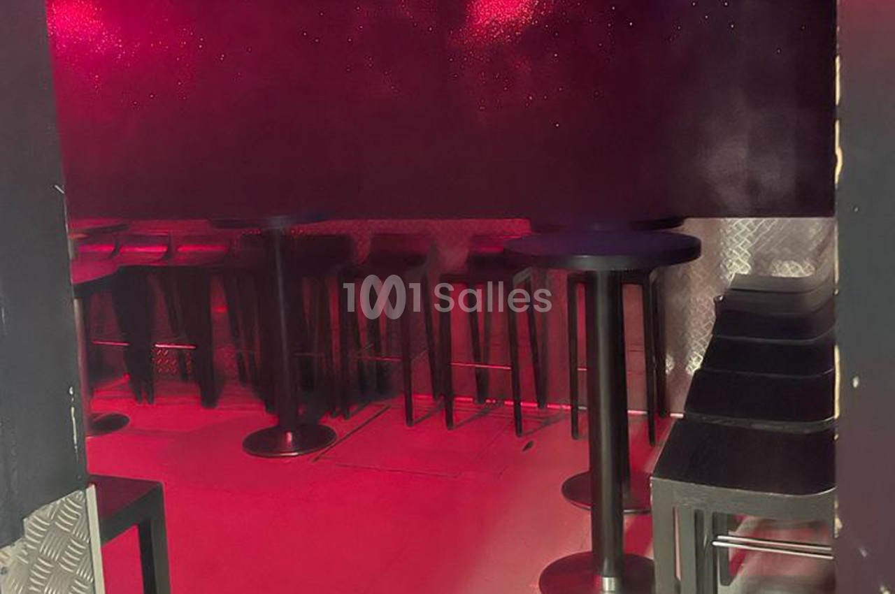 Salle sombre avec éclairage rouge, tables hautes noires et tabourets alignés le long des murs métalliques.
