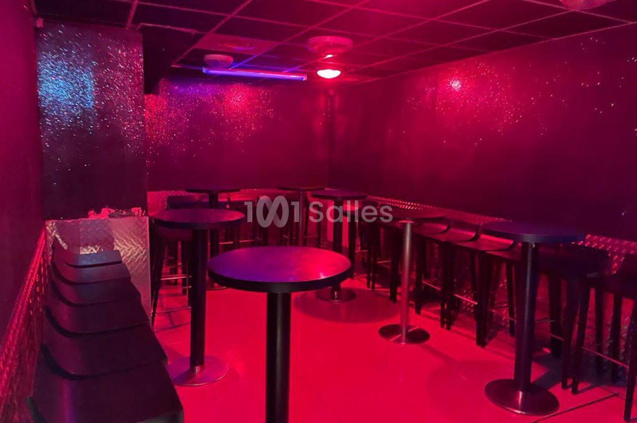 Salle sombre avec éclairage rouge, tables hautes noires, tabourets alignés et murs scintillants.