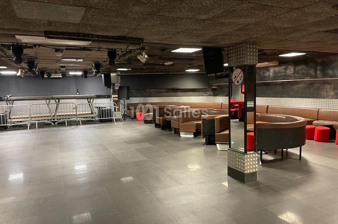 Salle vide avec sol en carrelage sombre, banquettes marron et rouges, éclairage tamisé et scène en arrière-plan.