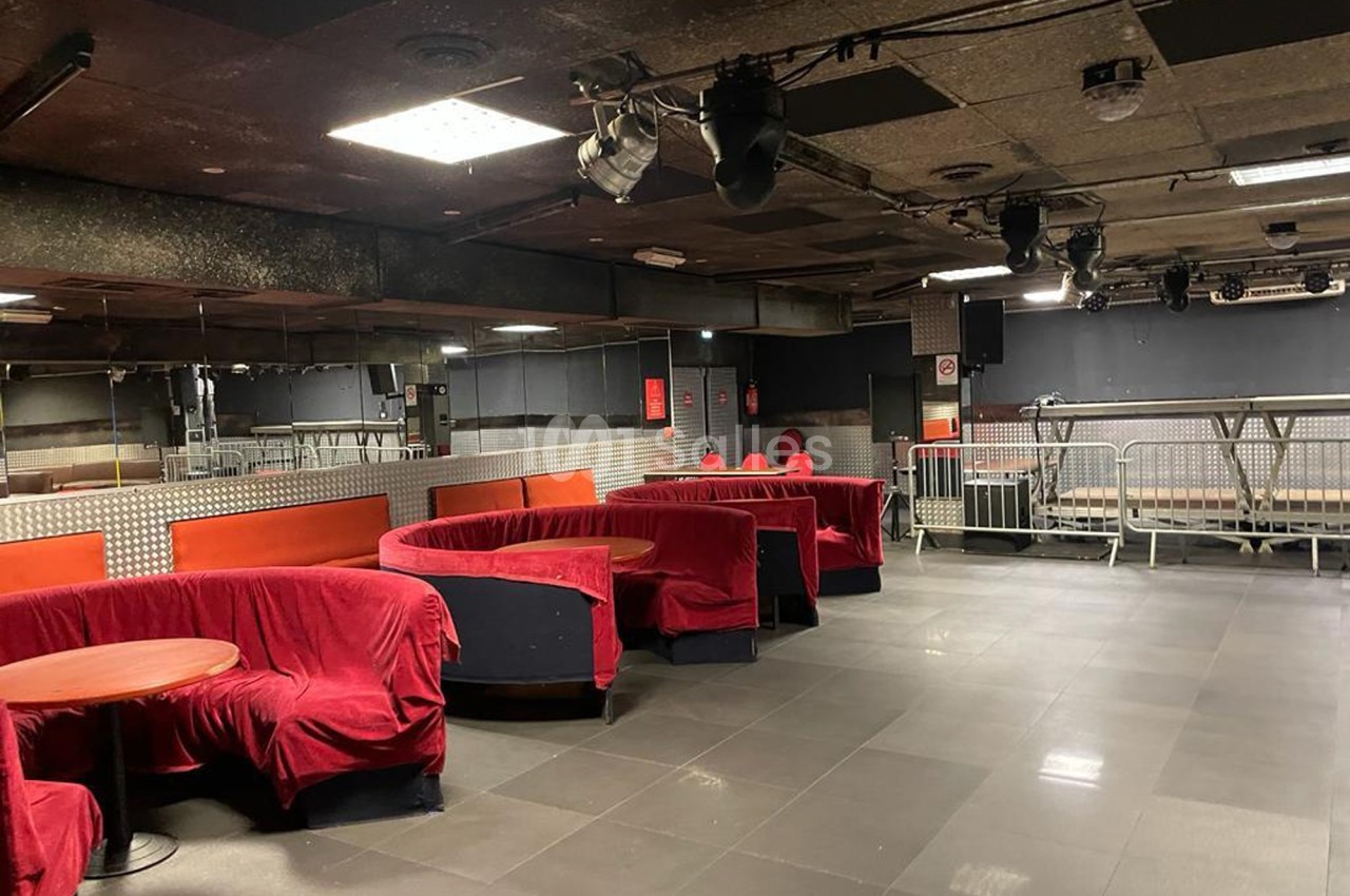 Salle vide avec banquettes rouges, tables rondes, sol carrelé gris et éclairage suspendu dans un espace industriel.