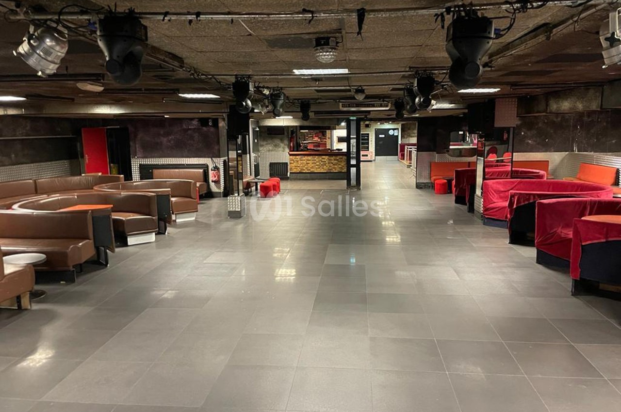 Salle vide avec canapés en cuir, tables basses et banquettes rouges, éclairage tamisé et sol en carrelage gris.
