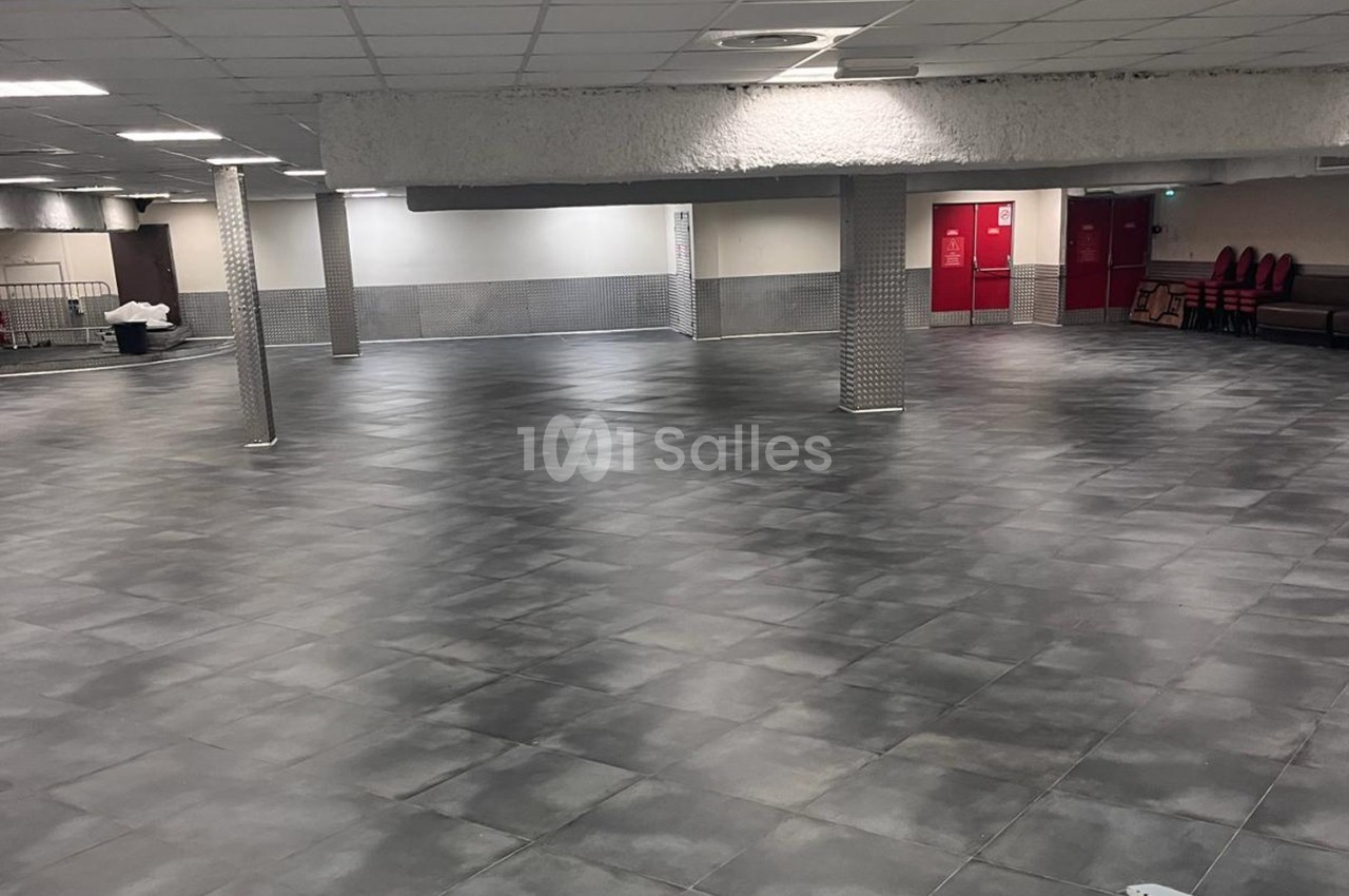 Salle vide avec sol en carrelage gris, piliers métalliques et murs clairs, éclairée par un plafond à dalles.