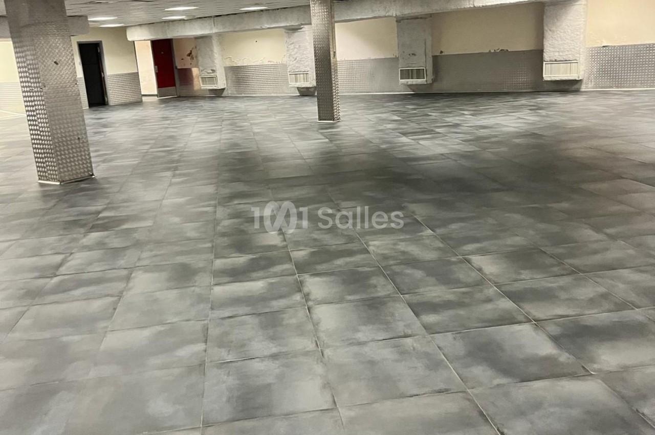 Salle vide avec un sol en carrelage gris, colonnes recouvertes de mosaïques et murs clairs partiellement carrelés.