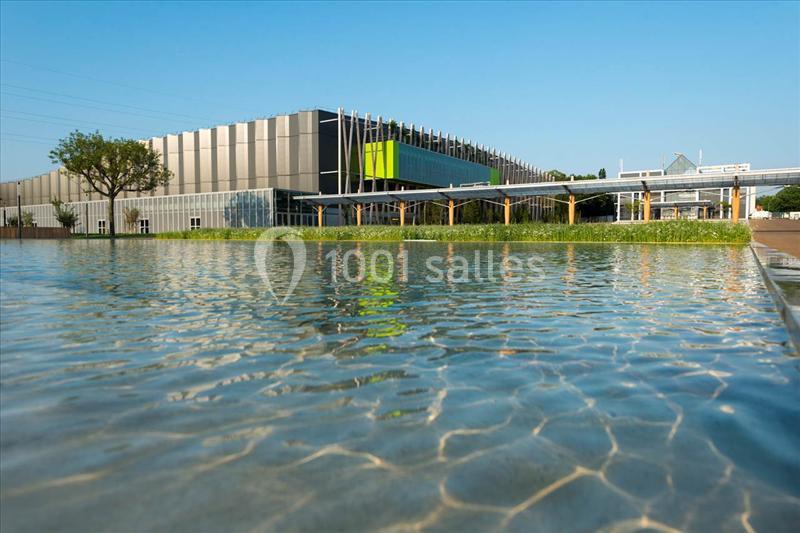 Vue d'un bâtiment moderne avec une façade vitrée, entouré d'un plan d'eau et de végétation sous un ciel dégagé.