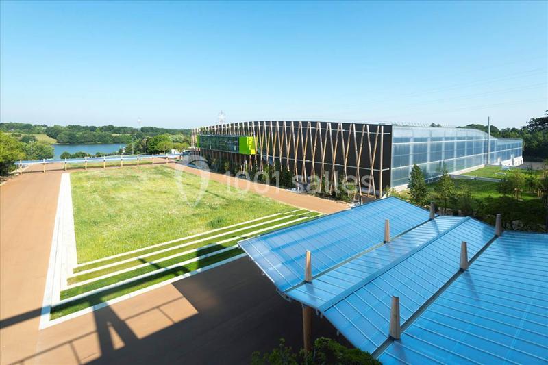 Vue d'un bâtiment moderne entouré de verdure, avec un toit en verre au premier plan et un lac en arrière-plan.