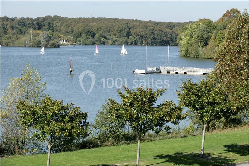 Plan d'eau bordé d'arbres avec des voiliers et une planche à voile, près d'un ponton en bois.