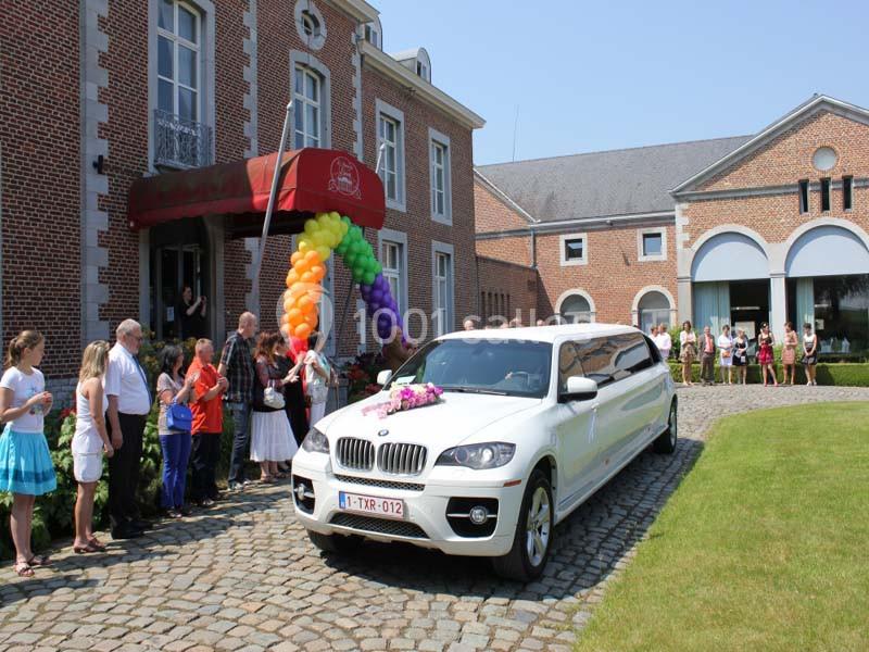 Une limousine blanche décorée de fleurs arrive devant un bâtiment en briques, entourée de spectateurs.
