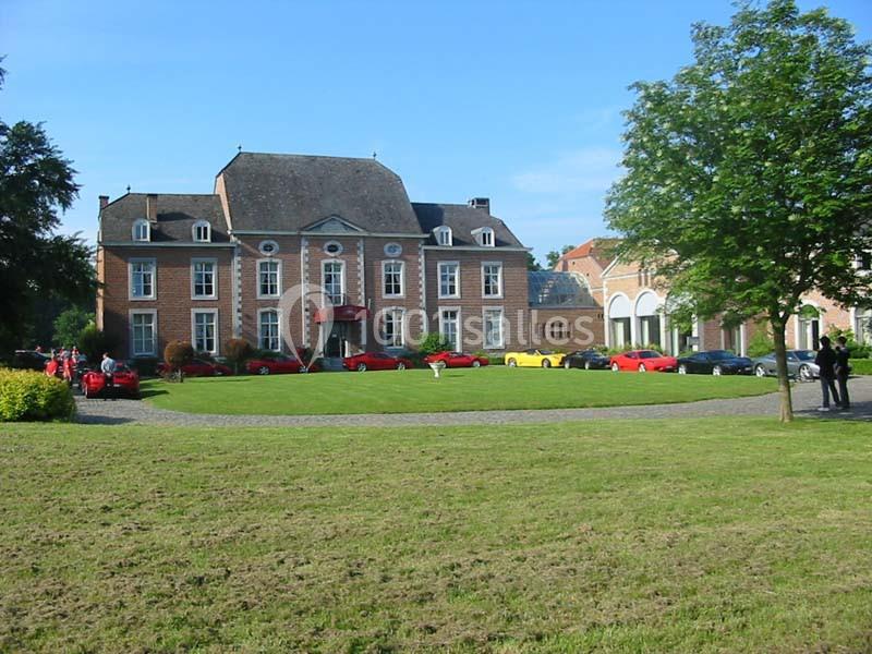 Façade d'un manoir en briques rouges entouré de pelouses, avec des voitures garées devant sur une allée pavée.