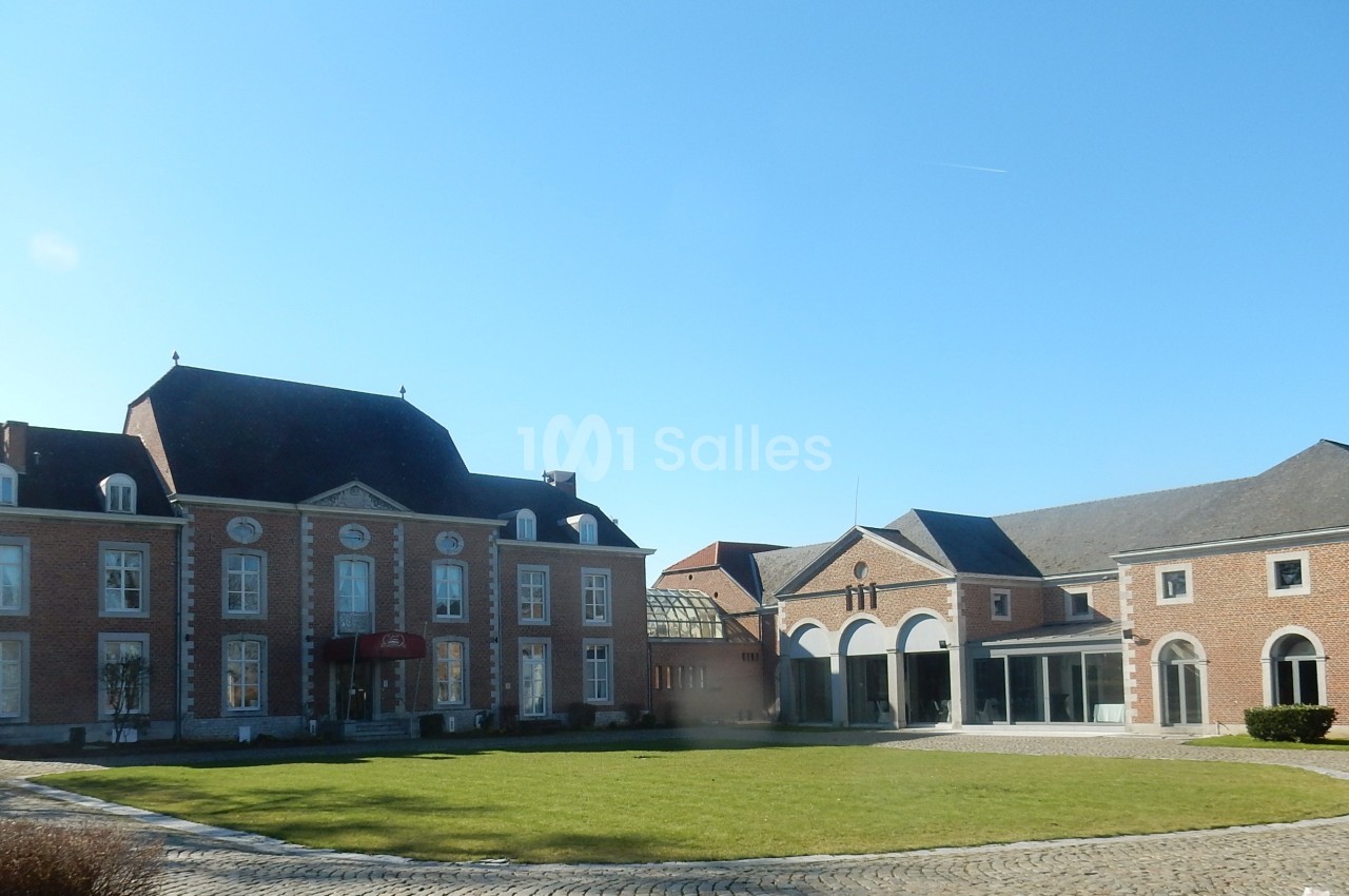 Bâtiment en briques rouges avec toit en ardoise, entouré d'une cour pavée et d'une pelouse sous un ciel dégagé.