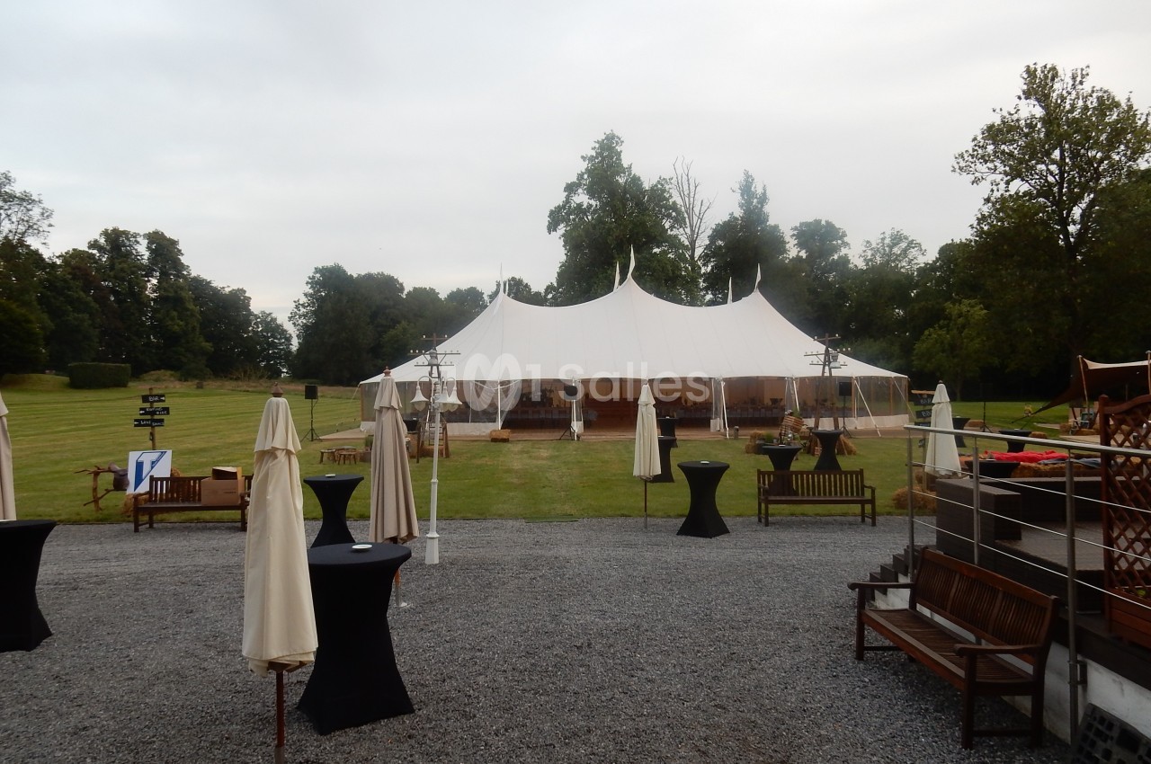 Grande tente blanche installée sur une pelouse entourée d'arbres, avec des tables et parasols disposés à l'extérieur.