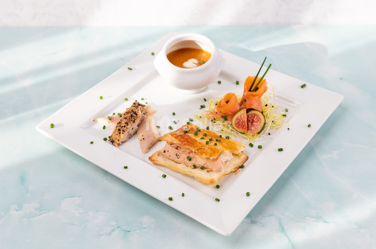 Assiette gastronomique avec feuilleté, poisson, saumon fumé, figue, germes de légumes et velouté en bol blanc.