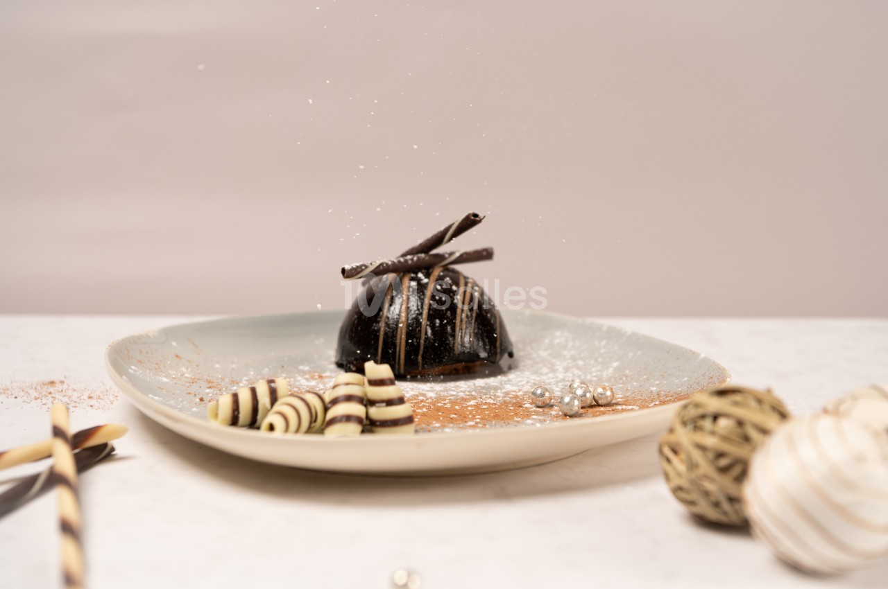 Dessert au chocolat décoré de spirales en chocolat et saupoudré de sucre glace, présenté sur une assiette beige.
