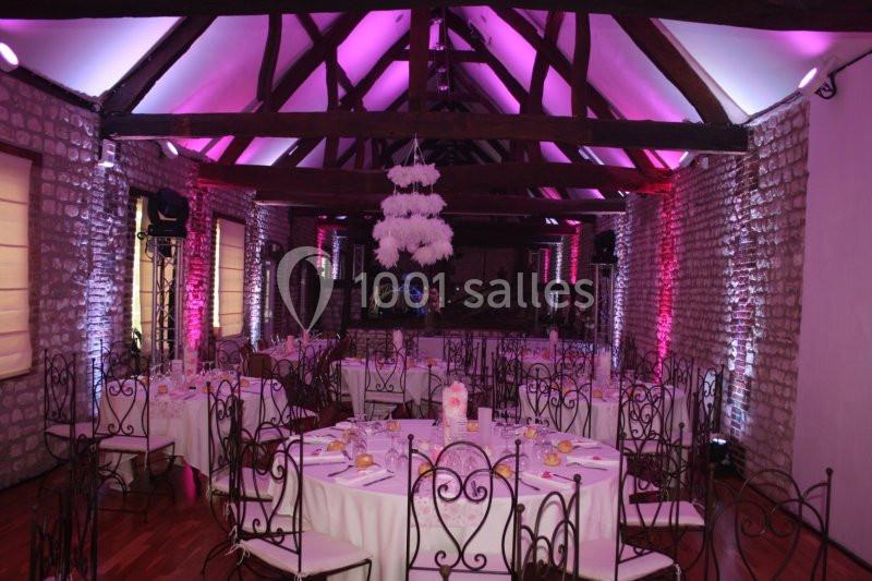 Salle de réception en pierre avec poutres apparentes, tables rondes dressées et éclairage rose.