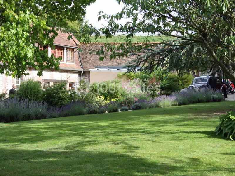 Jardin verdoyant avec pelouse, massifs de fleurs et arbres, devant une maison en pierre avec toit en tuiles.